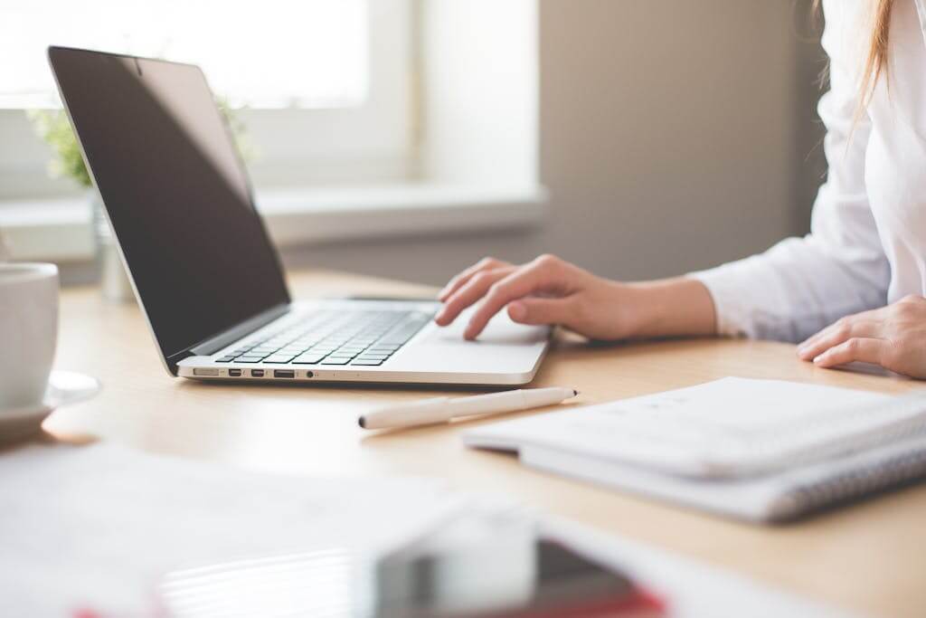 A businesswoman typing on a laptop at a bright indoor workspace with notebooks and a pen.